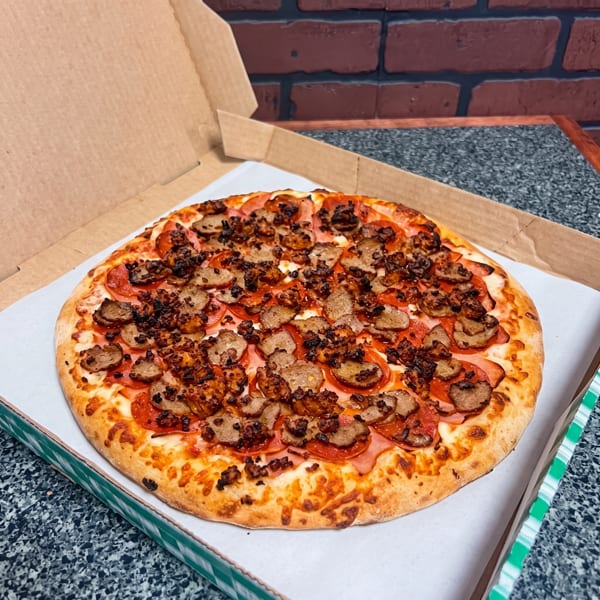 A freshly baked meat lover's pizza with pepperoni, ham, and sausage in an open delivery box.